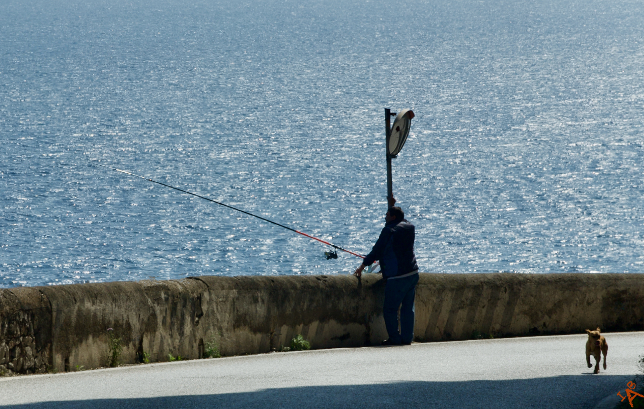 On the Amalfi Coast in Italy, a fisherman fishes while a dog walks by.